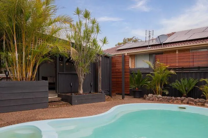 Una piscina exterior está junto a una casa revestida de madera y plantas tropicales.