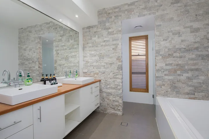 Un baño luminoso con un tocador largo y una bañera junto a paredes con acabado de piedra.