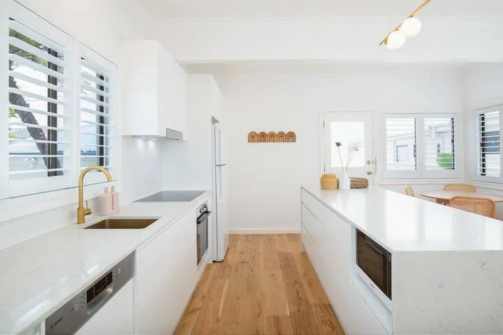 Una cocina blanca y luminosa tiene una isla larga y suelo de madera.