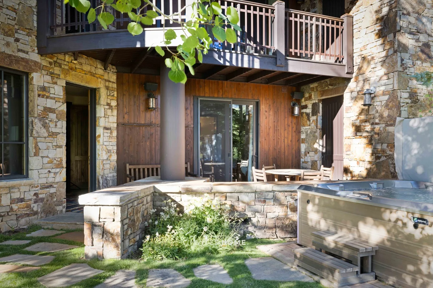 Un patio de piedra con asientos al aire libre y una bañera de hidromasaje junto a una casa revestida de madera.