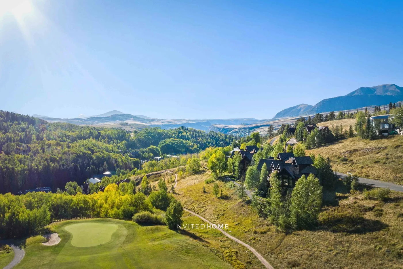 Un campo de golf y un sendero serpenteante se encuentran entre colinas boscosas y montañas lejanas.