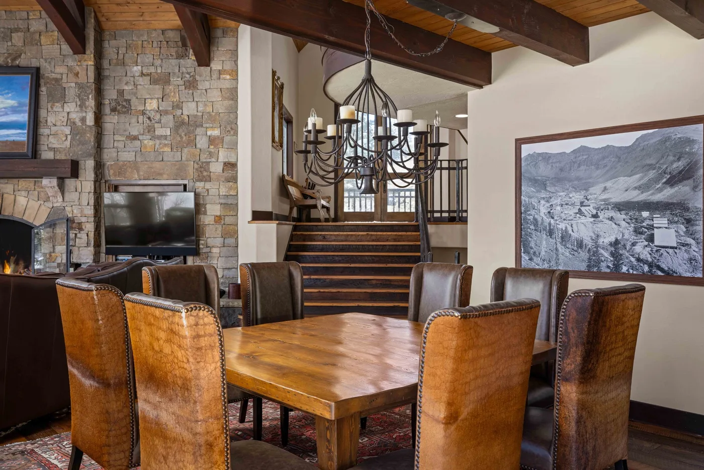 Comedor con una mesa de madera, asientos tapizados y una lámpara de araña bajo vigas vistas.