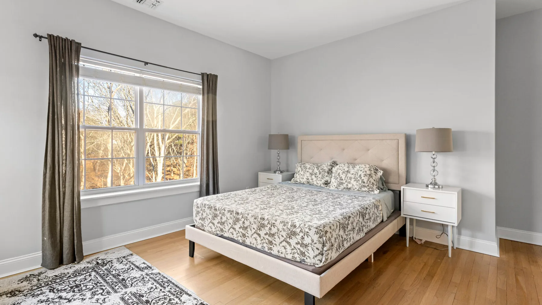 Un dormitorio luminoso con una cama, mesitas de noche y una gran ventana con cortinas.