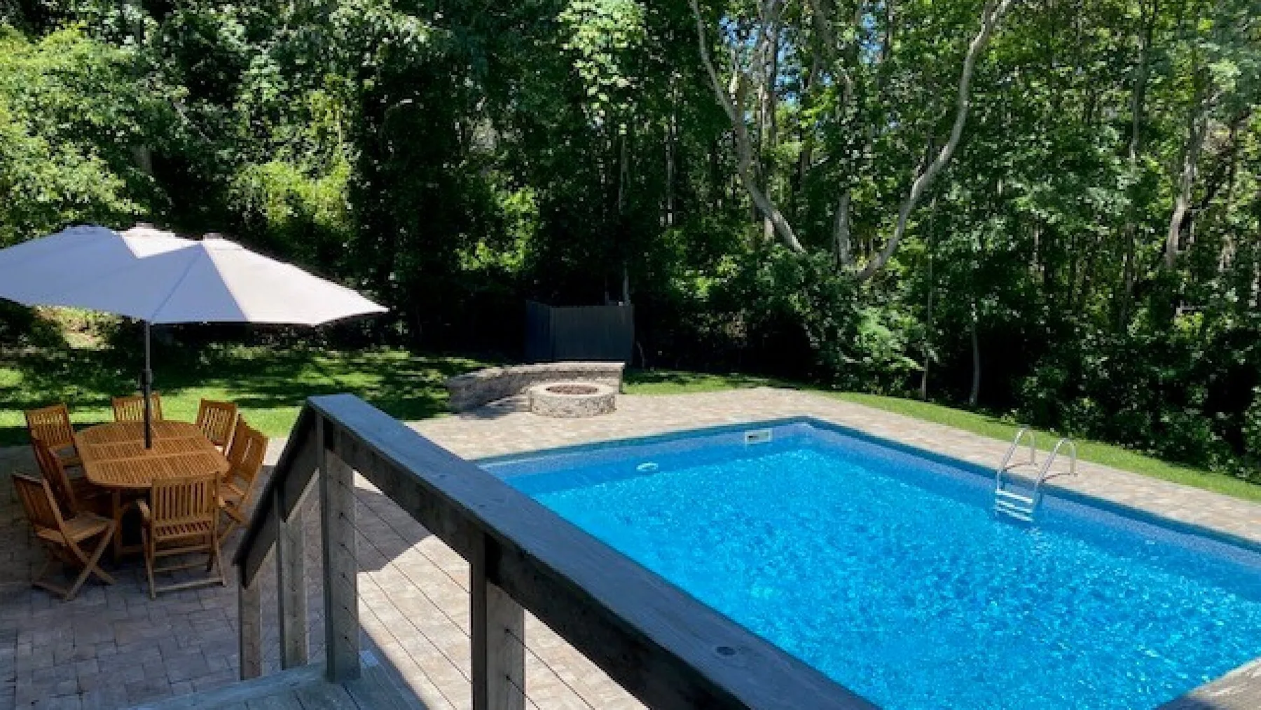 Una piscina al aire libre junto a una zona de comedor en el patio, rodeada de árboles.