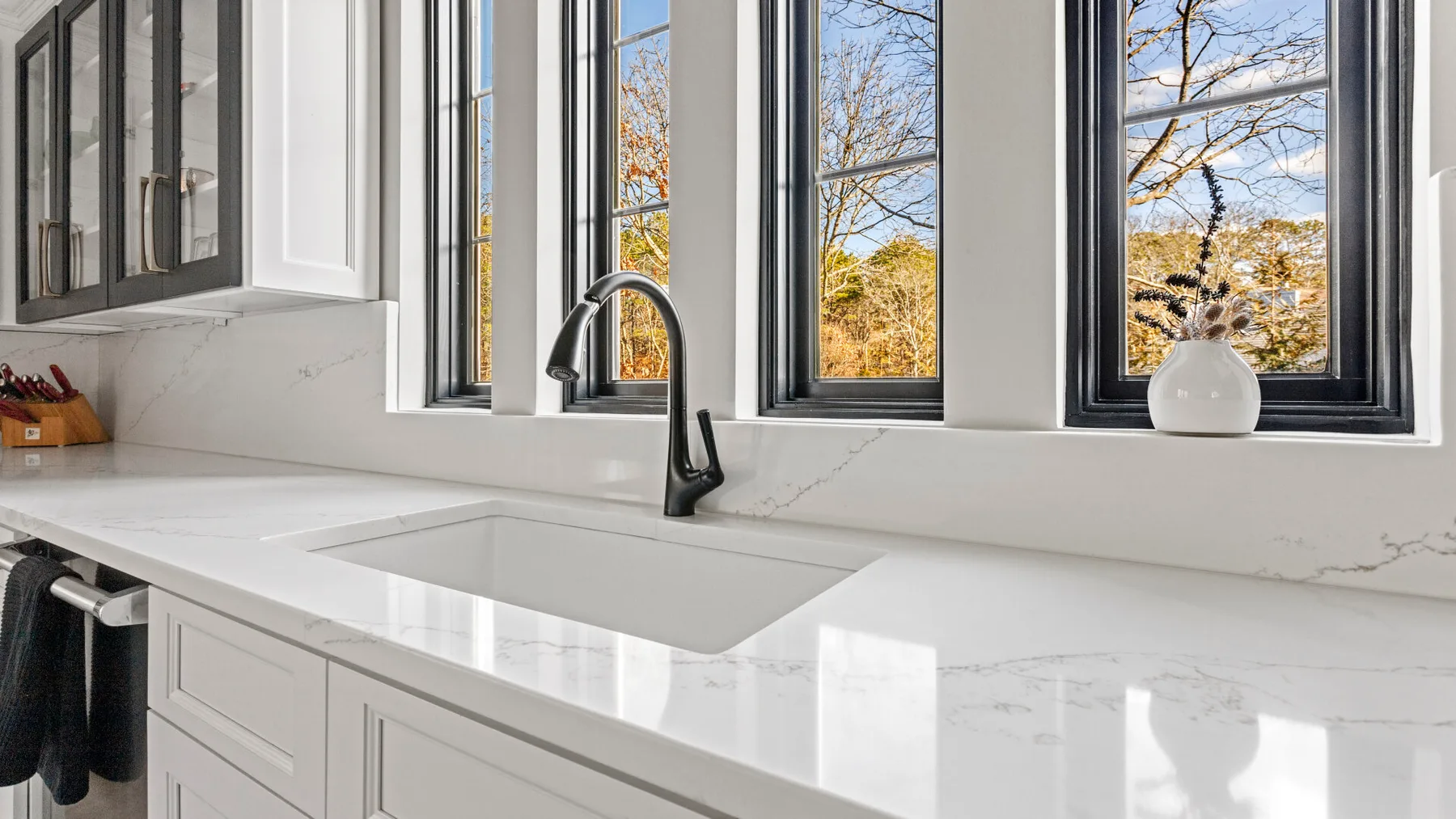 Una cocina luminosa con un fregadero blanco bajo grandes ventanas.