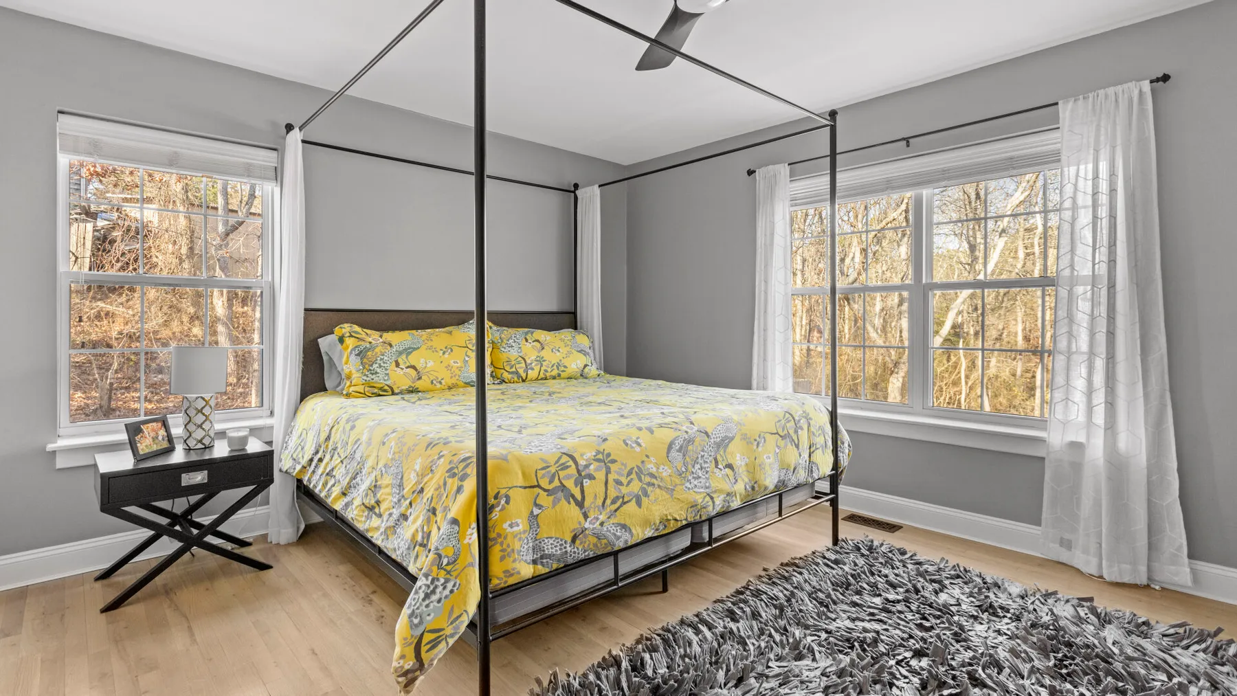 Un dormitorio con una cama cubierta con ropa de cama amarilla con flores, junto a grandes ventanas.