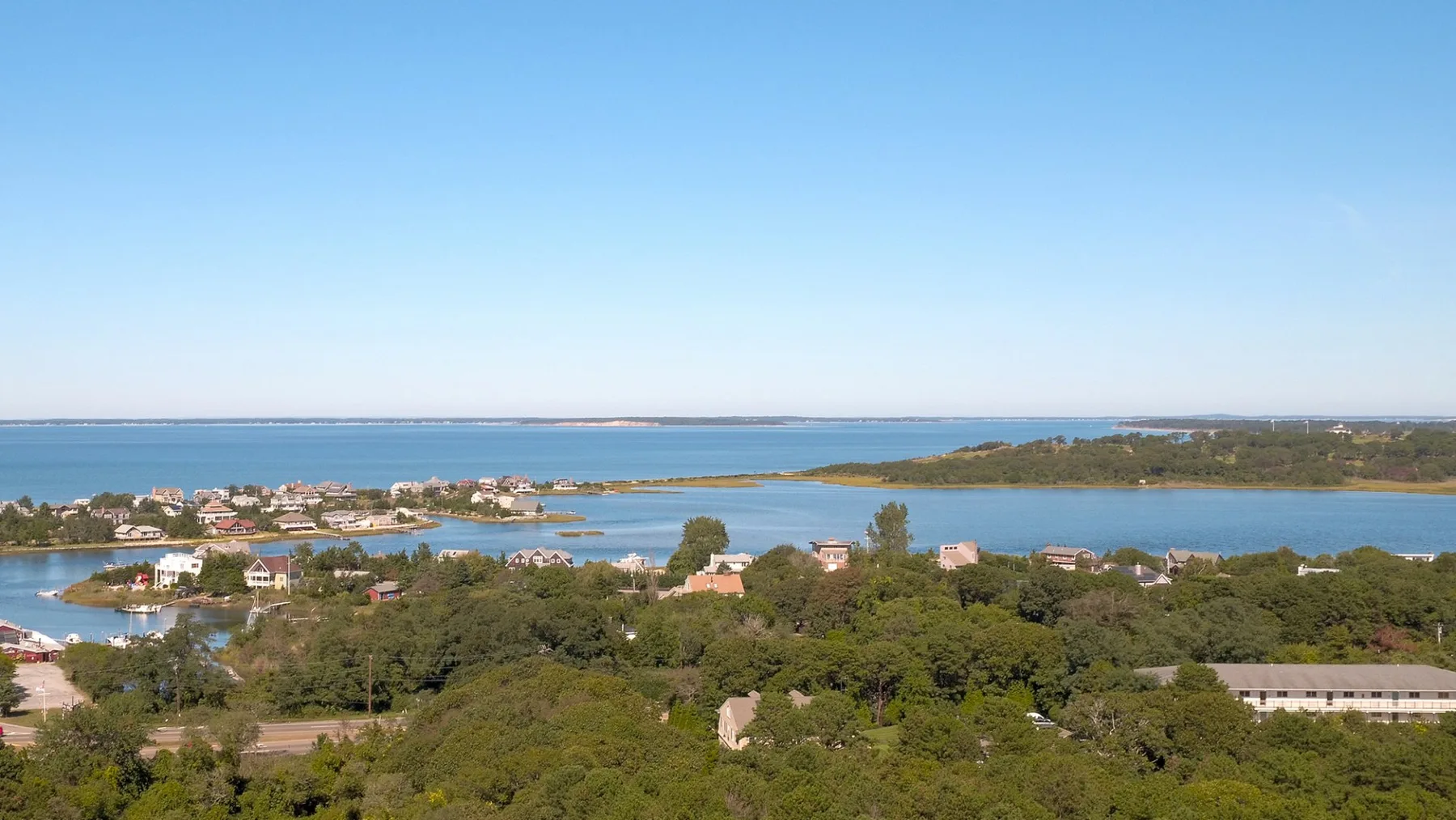 Un amplio paisaje costero muestra una bahía azul tranquila detrás de árboles verdes y edificios dispersos.