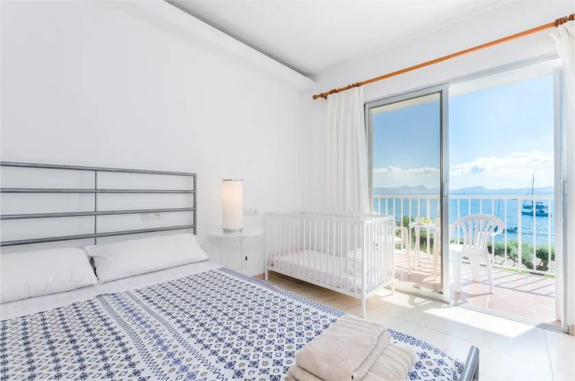A bright bedroom with a bed and a crib beside open doors overlooking the sea.