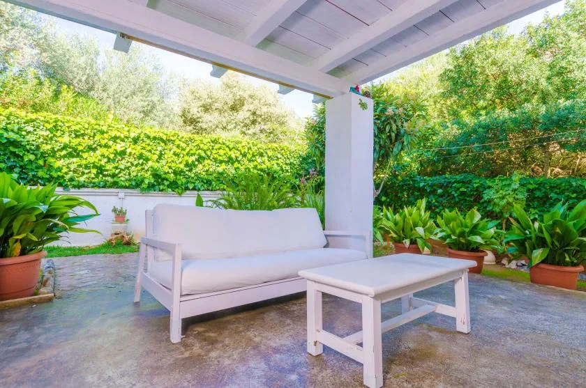 A covered patio with white seating surrounded by leafy plants.