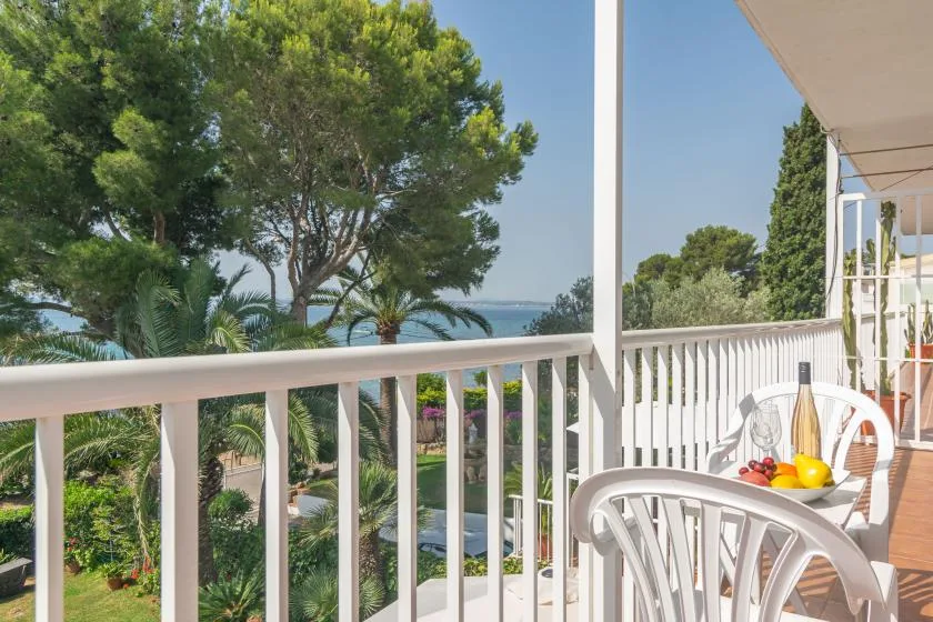 A balcony with white railings and seating overlooks leafy gardens and the sea.