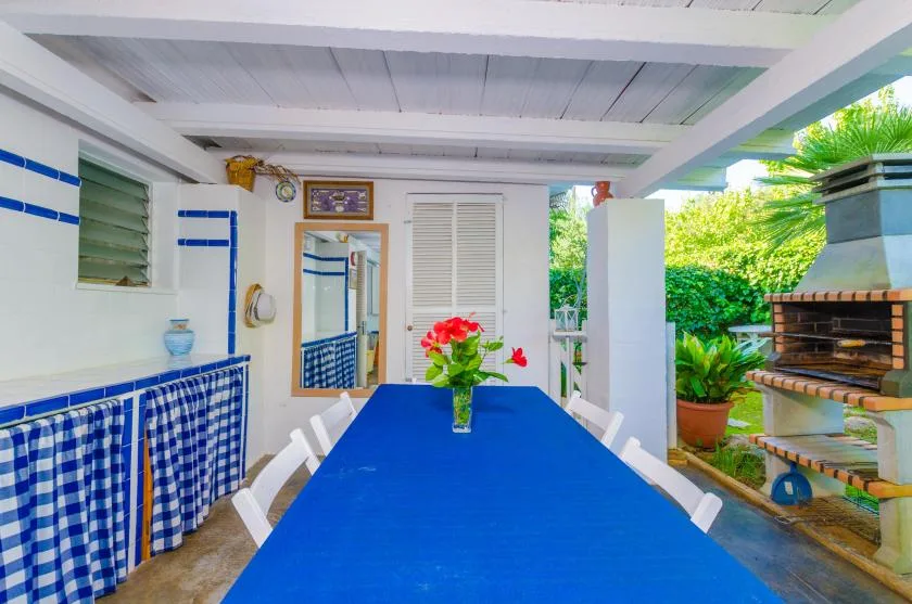 A covered patio dining area has a long blue table beside an outdoor grill.