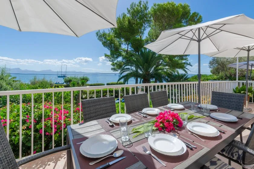 A terrace dining table with place settings and umbrellas overlooks a garden and water.