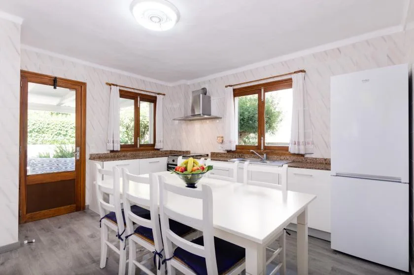 A bright kitchen with a white dining table, chairs, and windows overlooking greenery.