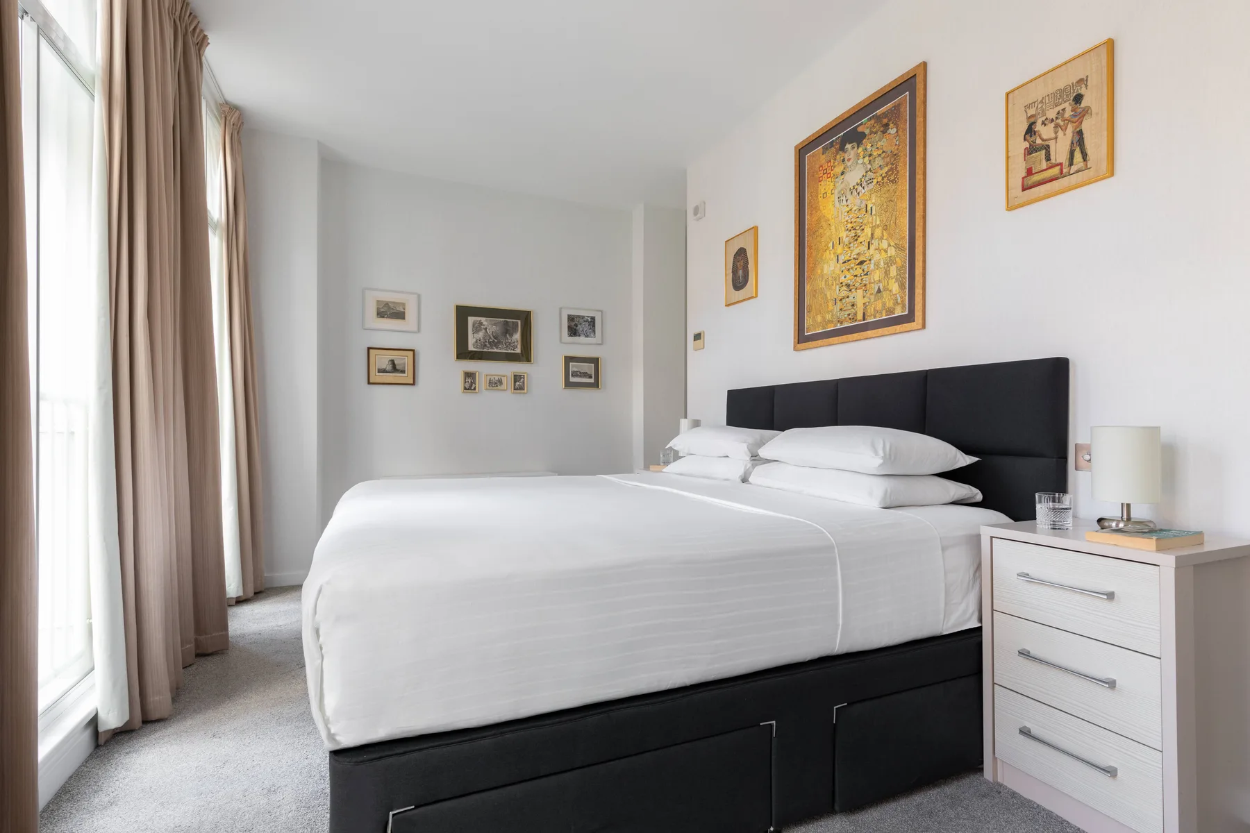 A bedroom with a neatly made bed, framed artwork, and a bedside chest.