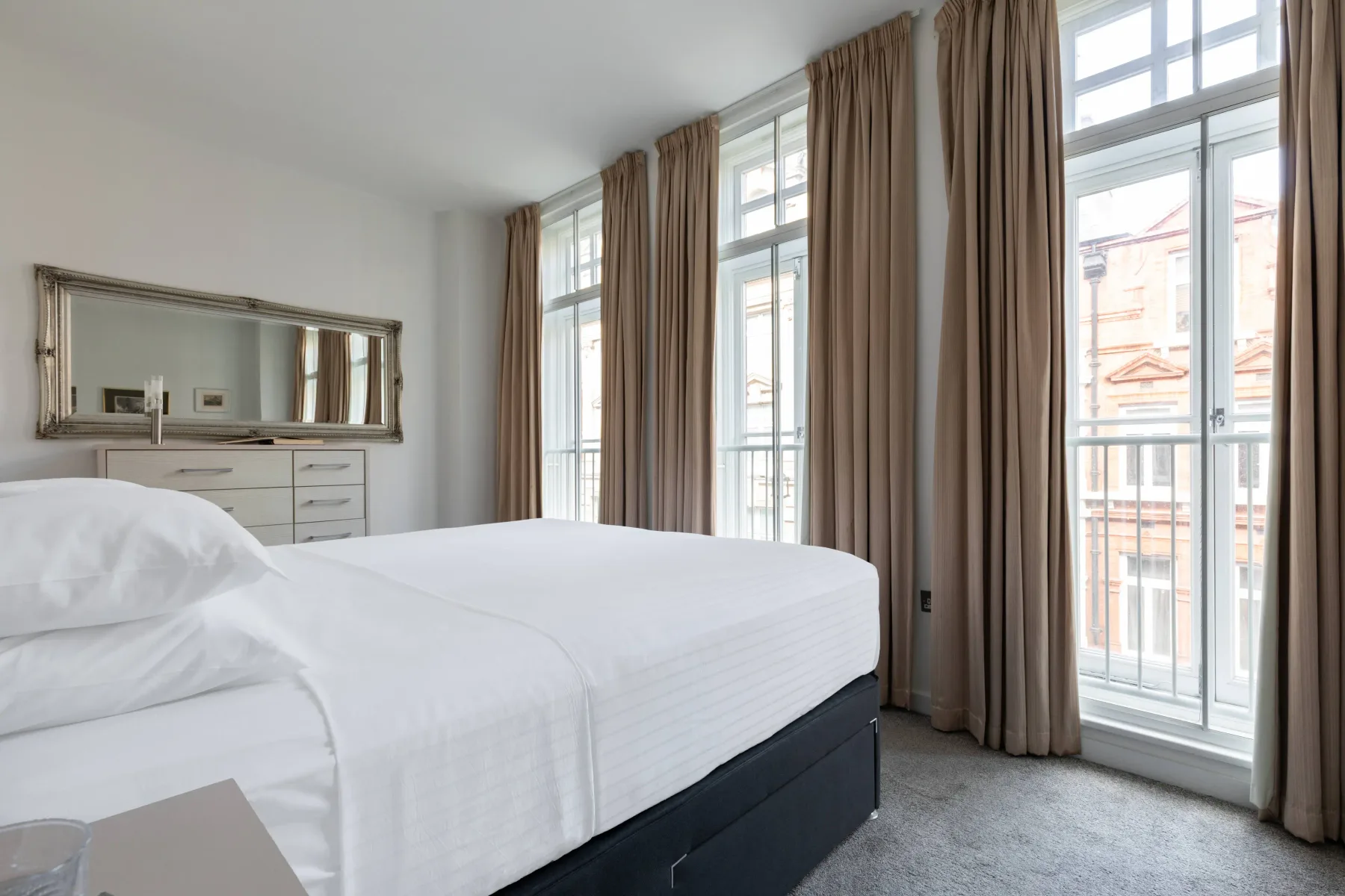 A bright bedroom with a neatly made bed beside tall windows.