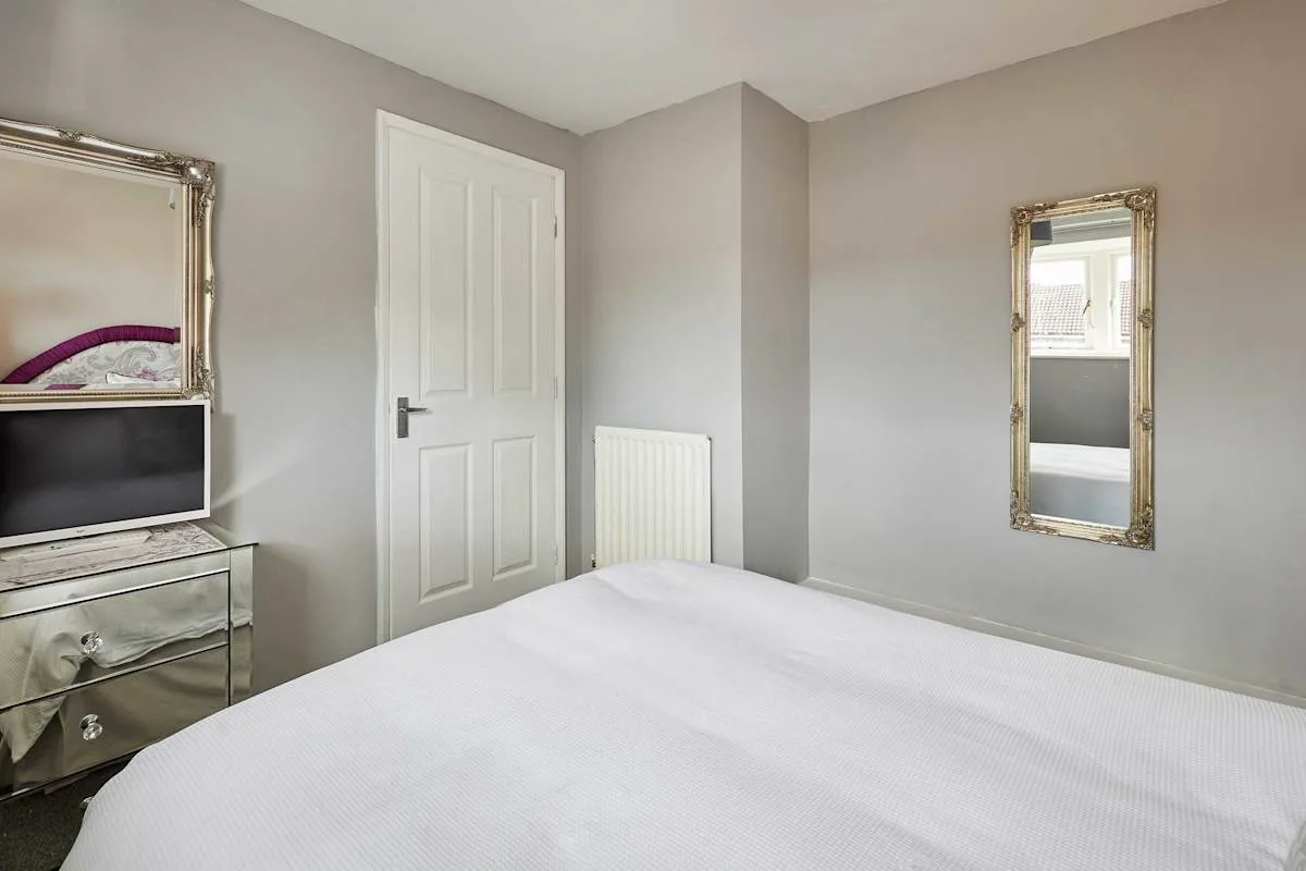 Un dormitorio con una cama blanca, un televisor sobre una cómoda y un espejo de pared.