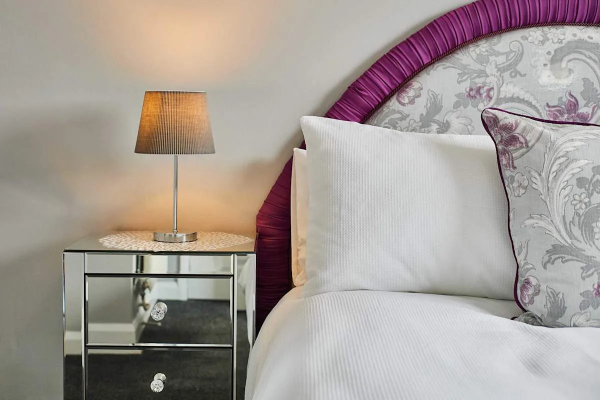 Un dormitorio con una cama blanca, cojines estampados y una lámpara de noche.