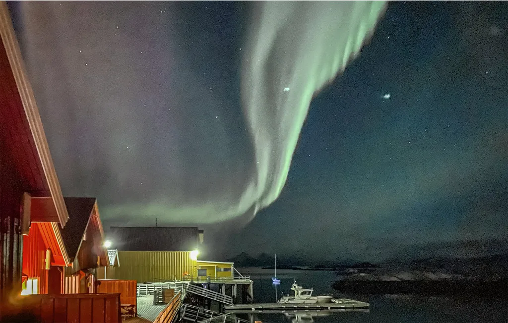 Un muelle junto al agua y cabañas rojas aparecen bajo auroras verdes en el cielo nocturno.