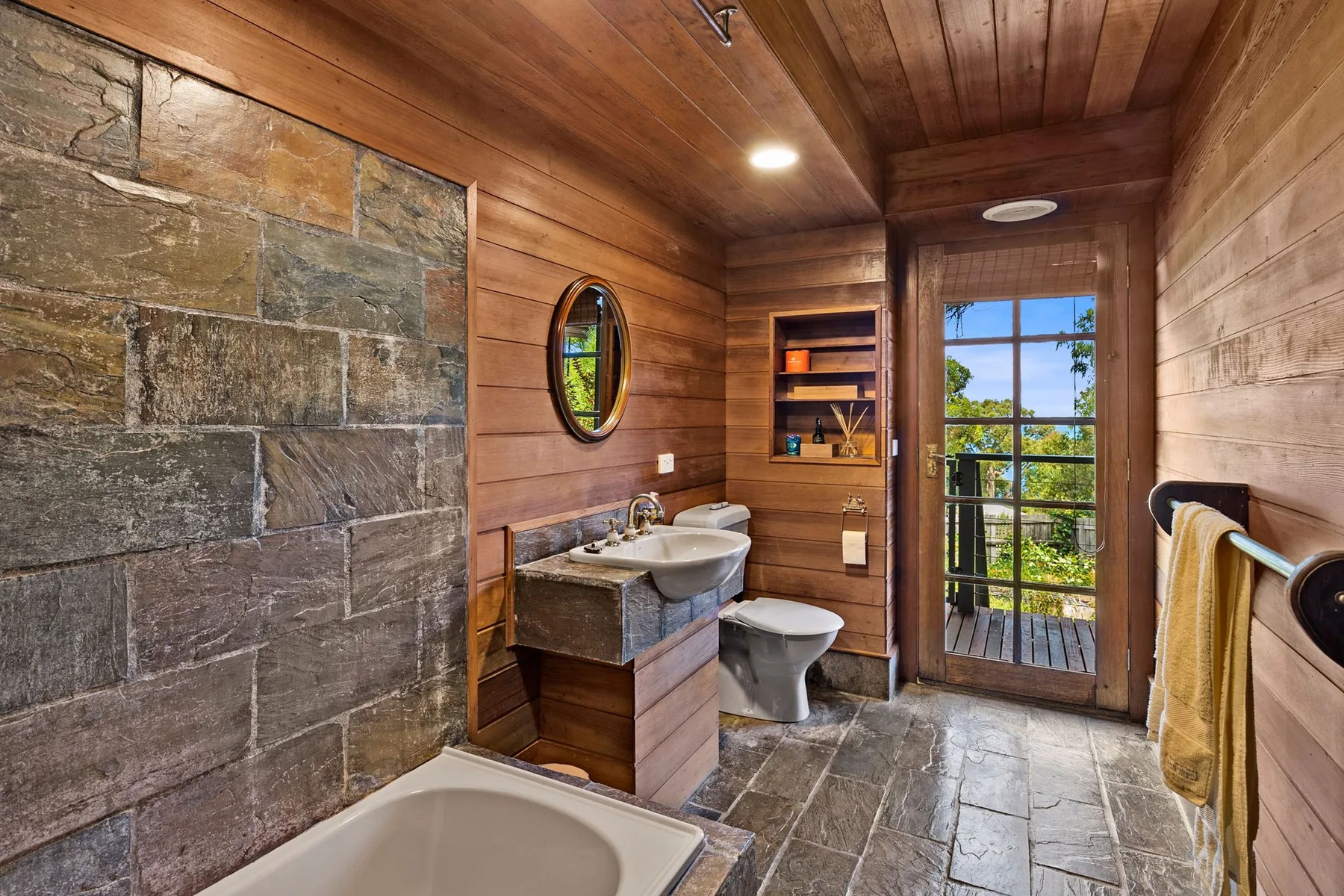 Un baño revestido de madera con bañera, lavabo y vista a través de una puerta de vidrio.