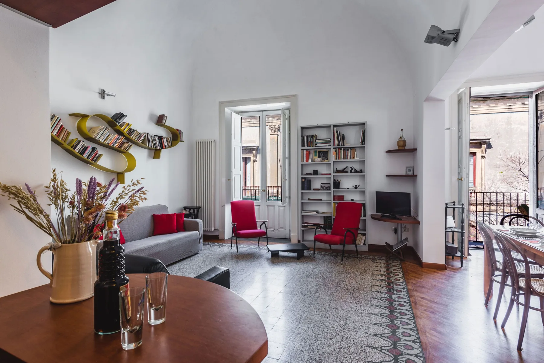 A bright living room with seating, bookshelves, and tall doors opening onto a balcony.