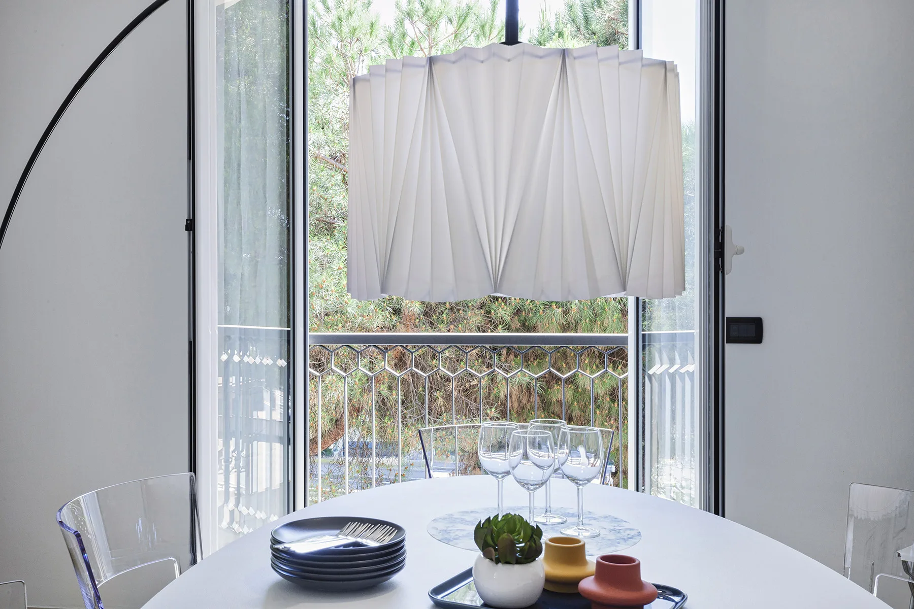 A dining table with place settings sits beside a tall window overlooking greenery.
