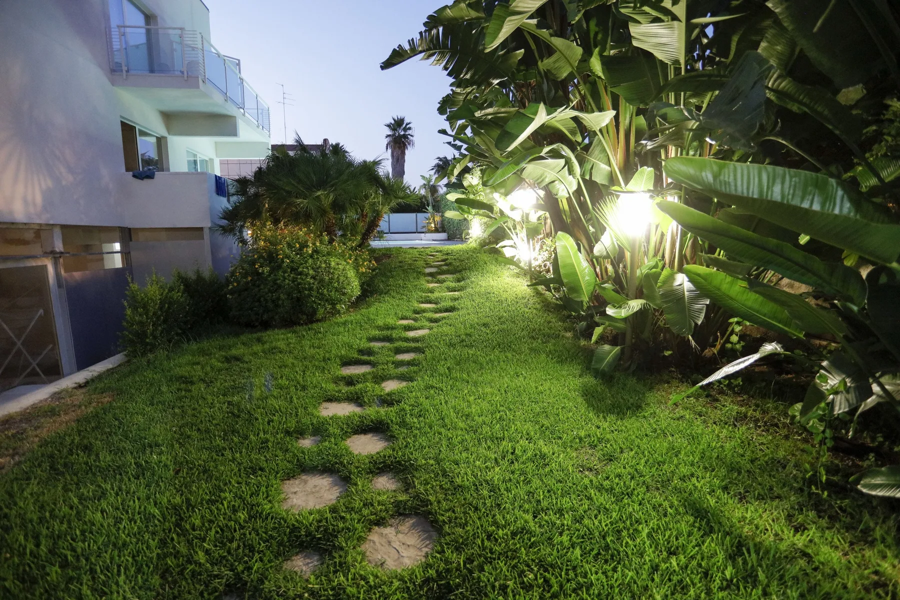 Un sendero de jardín atraviesa el césped entre plantas frondosas junto a un edificio moderno.