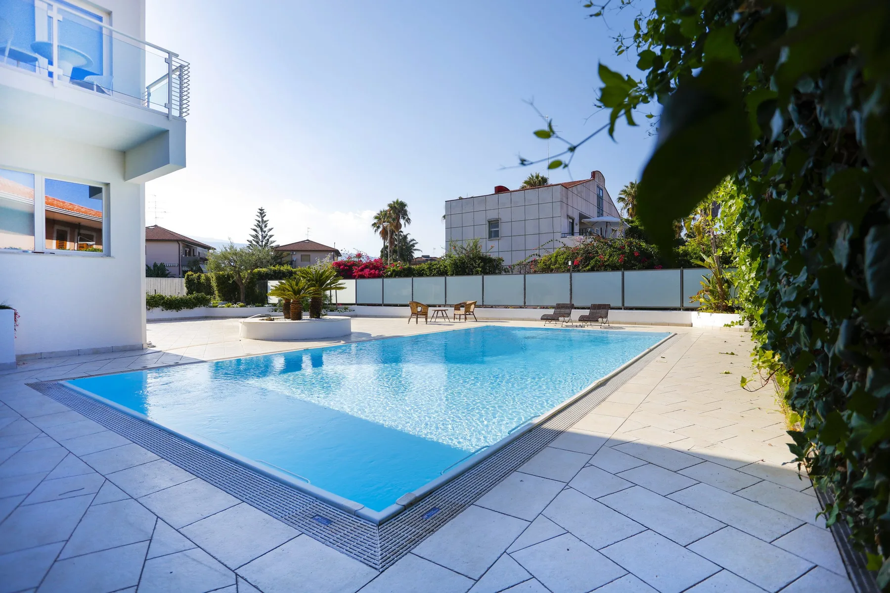 Una piscina al aire libre está rodeada de pavimento claro y vegetación.