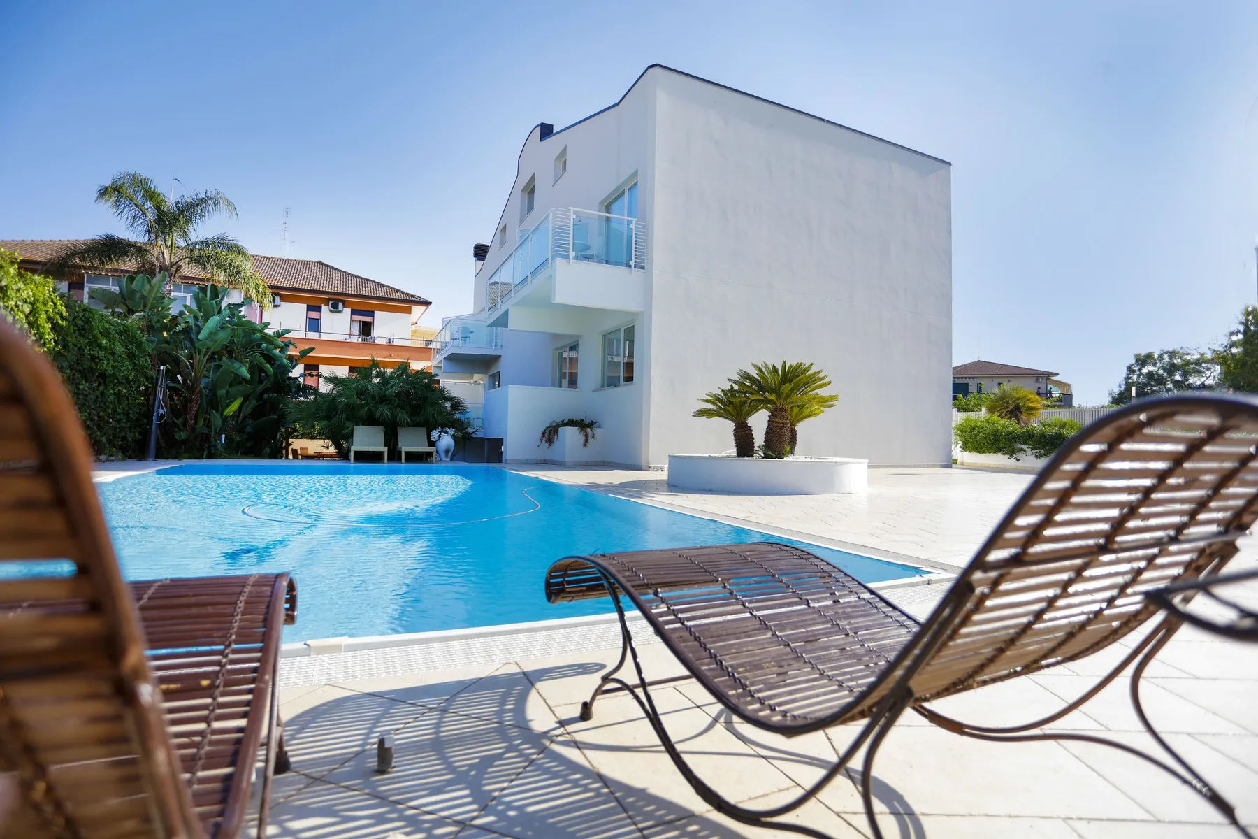 Una piscina al aire libre junto a un edificio blanco moderno, con asientos en la terraza.