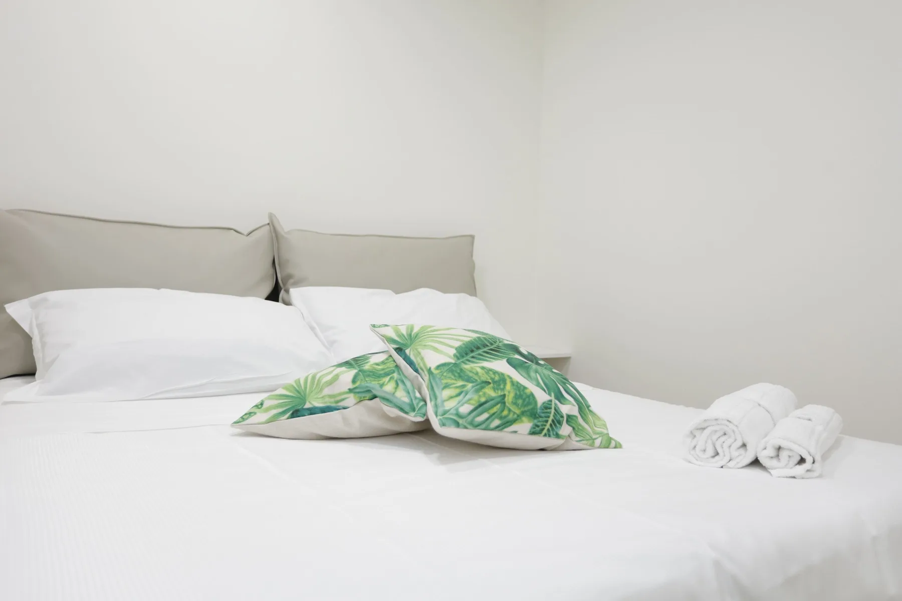 Una cama con ropa de cama blanca, cojines con hojas verdes y toallas enrolladas.