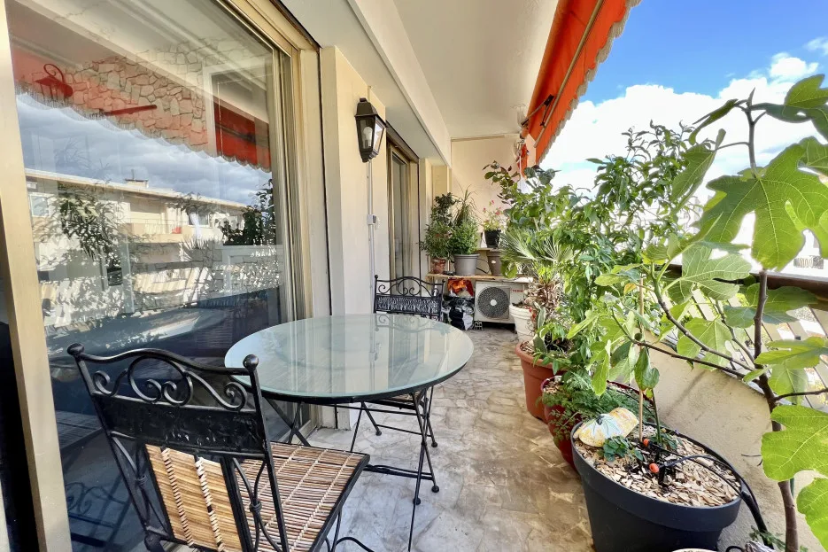 A narrow balcony with a glass-top table, metal seating, and potted plants.
