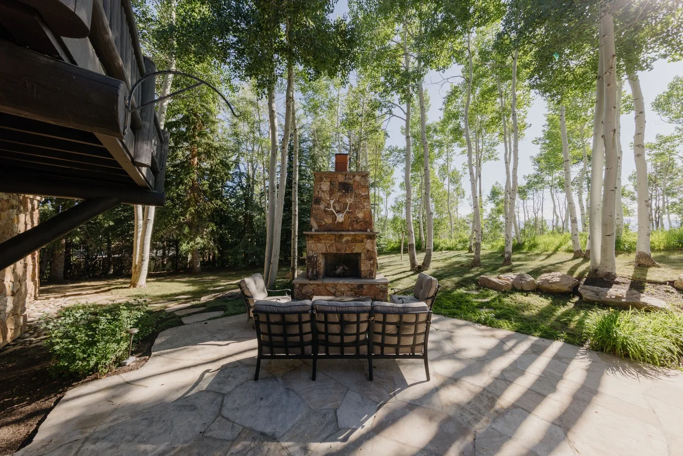 Un patio exterior con asientos acolchados frente a una chimenea de piedra entre árboles altos.