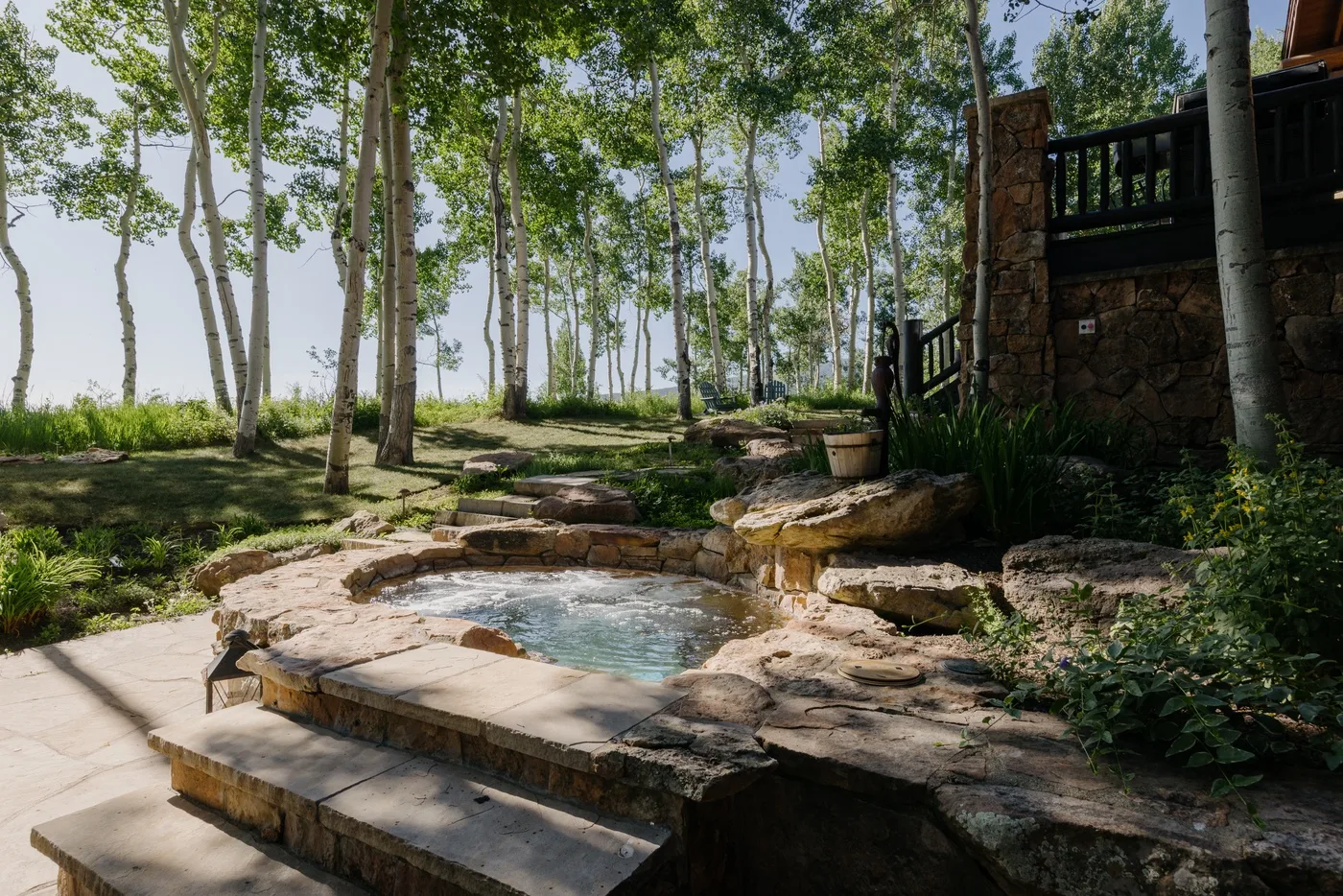 Una piscina con borde de piedra está junto a un jardín arbolado y una terraza elevada.