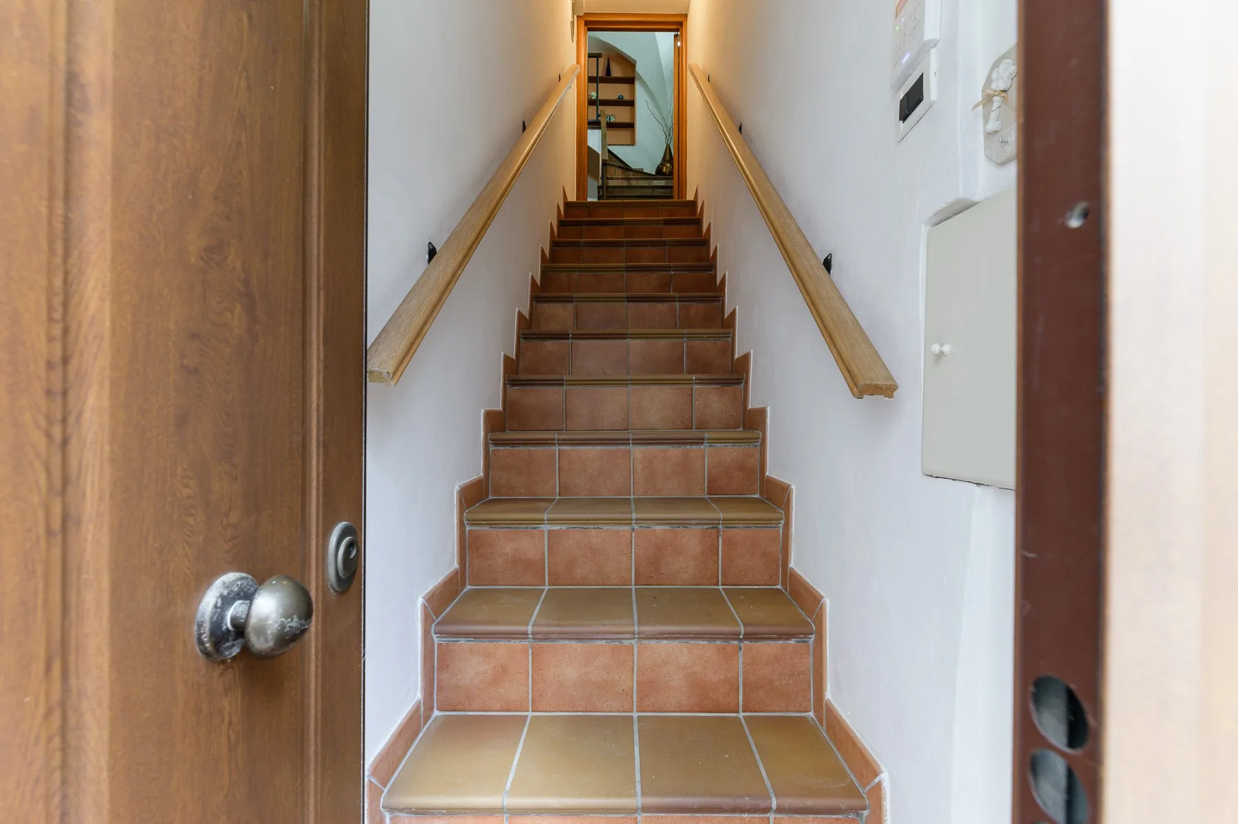Una escalera de azulejos sube entre paredes blancas con pasamanos de madera.