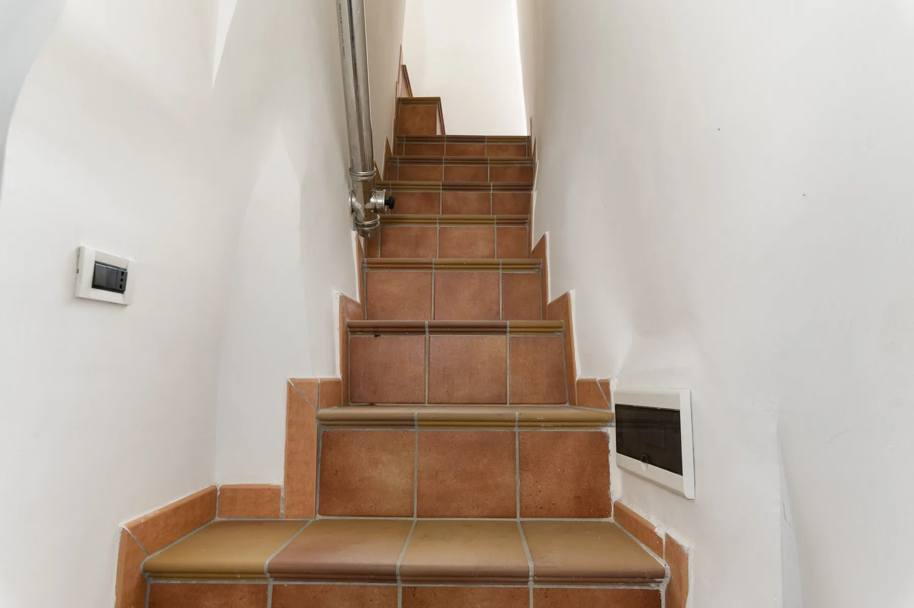 Una escalera estrecha de azulejos sube entre paredes blancas.