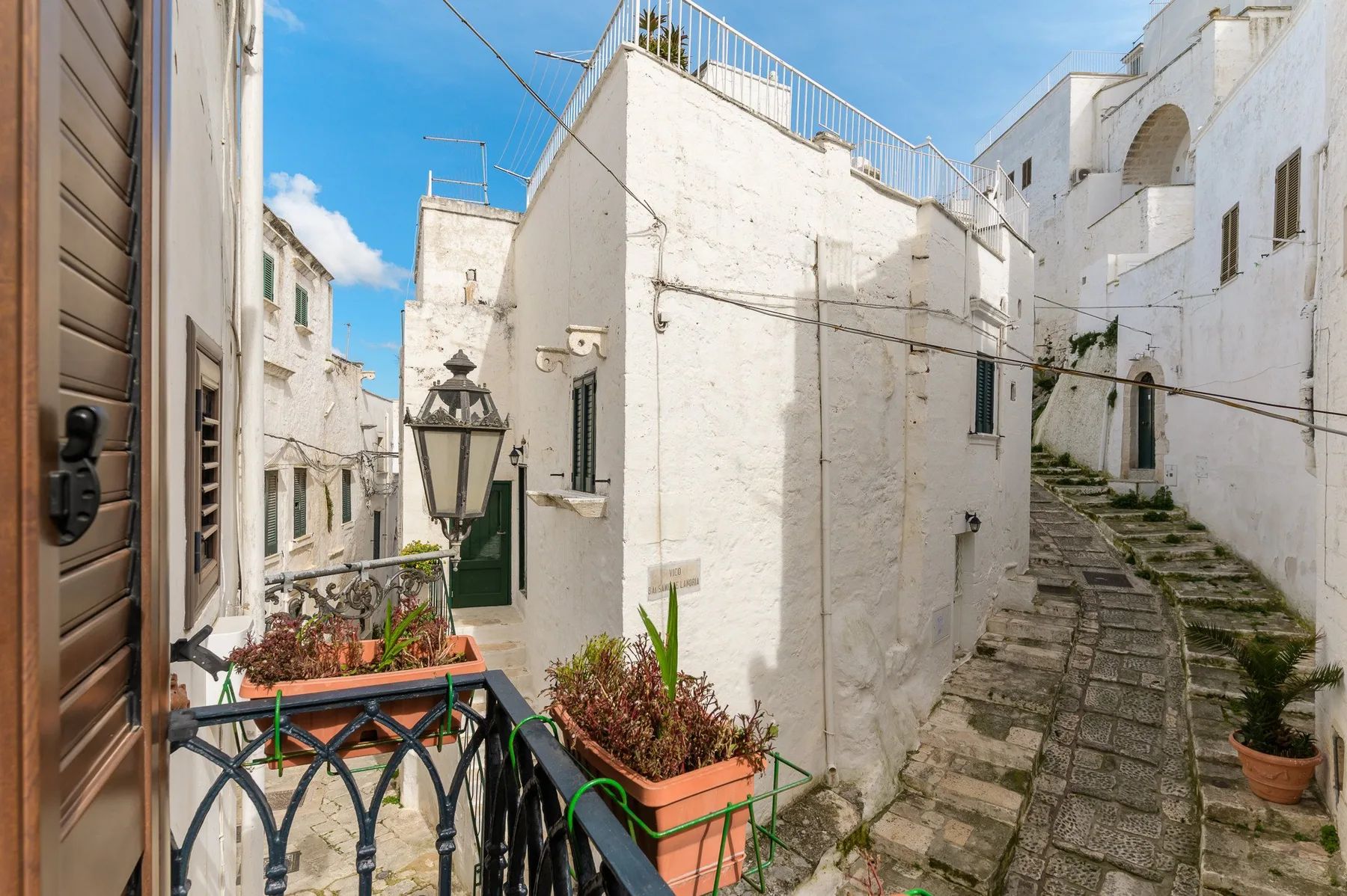 Un estrecho callejón de piedra pasa entre edificios blancos, con plantas en macetas a lo largo.