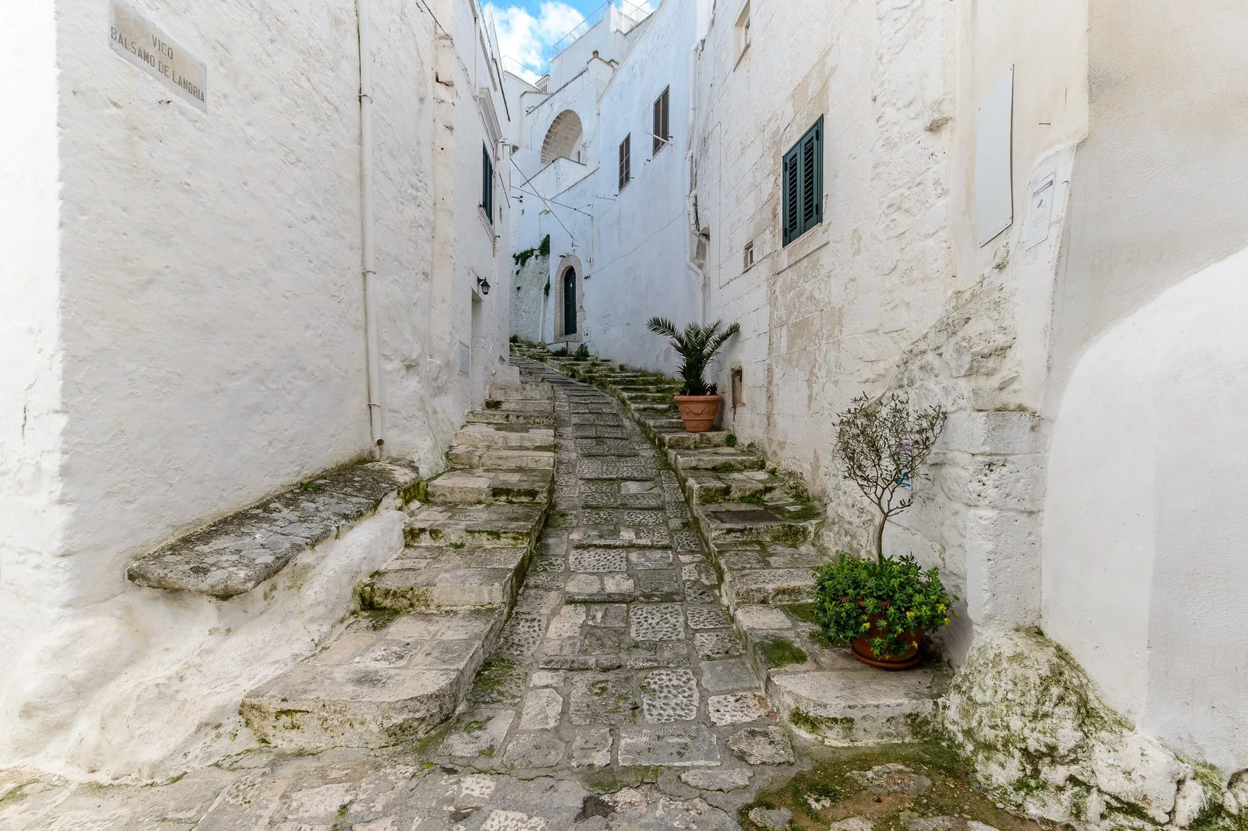 Una estrecha calle de piedra sube entre edificios encalados, con plantas en macetas junto al camino.