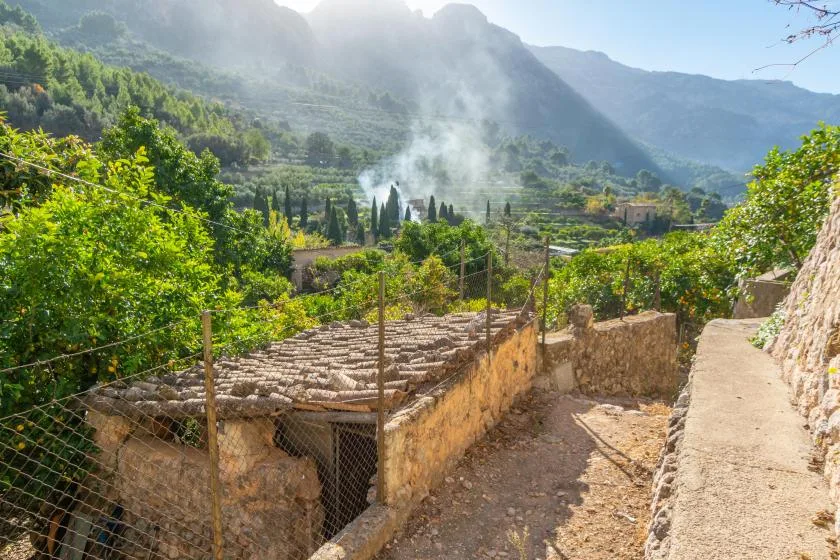 Un sendero de piedra pasa junto a edificios en la ladera, con terrazas verdes y montañas al fondo.