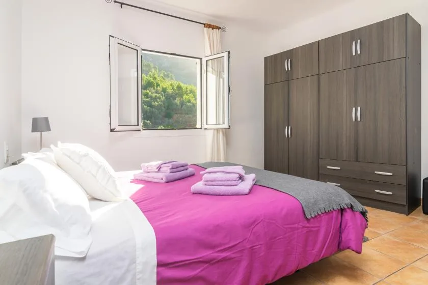 Un dormitorio con una cama cubierta con ropa de cama rosa junto a una ventana abierta.