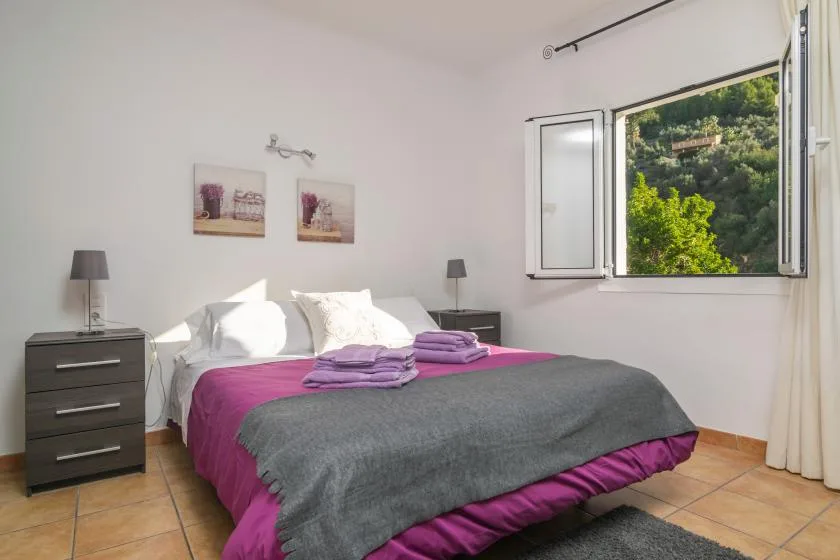 Un dormitorio con una cama vestida de morado y gris junto a una ventana abierta.