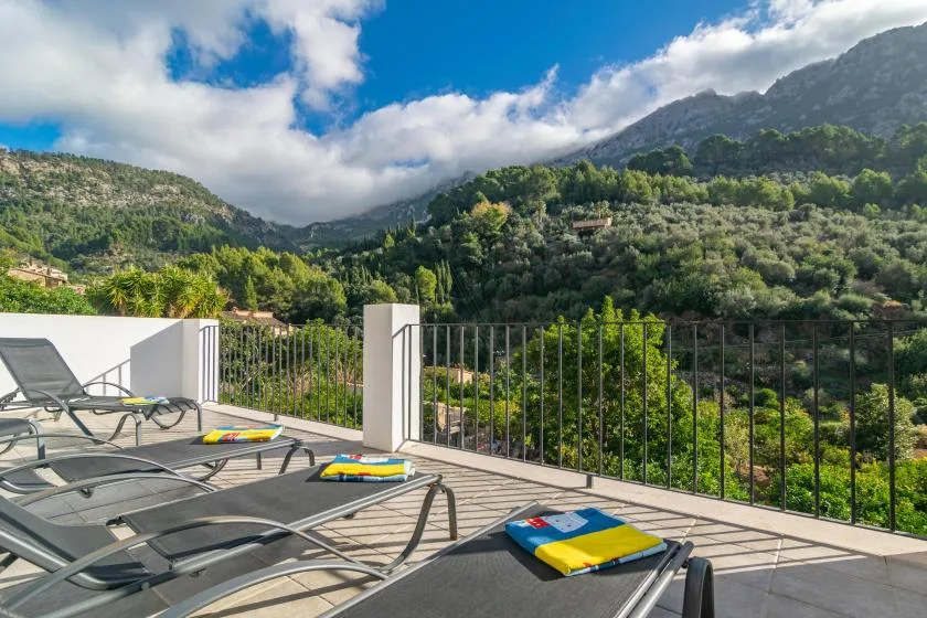 A sunlit terrace with lounge seating overlooks a green mountain valley.