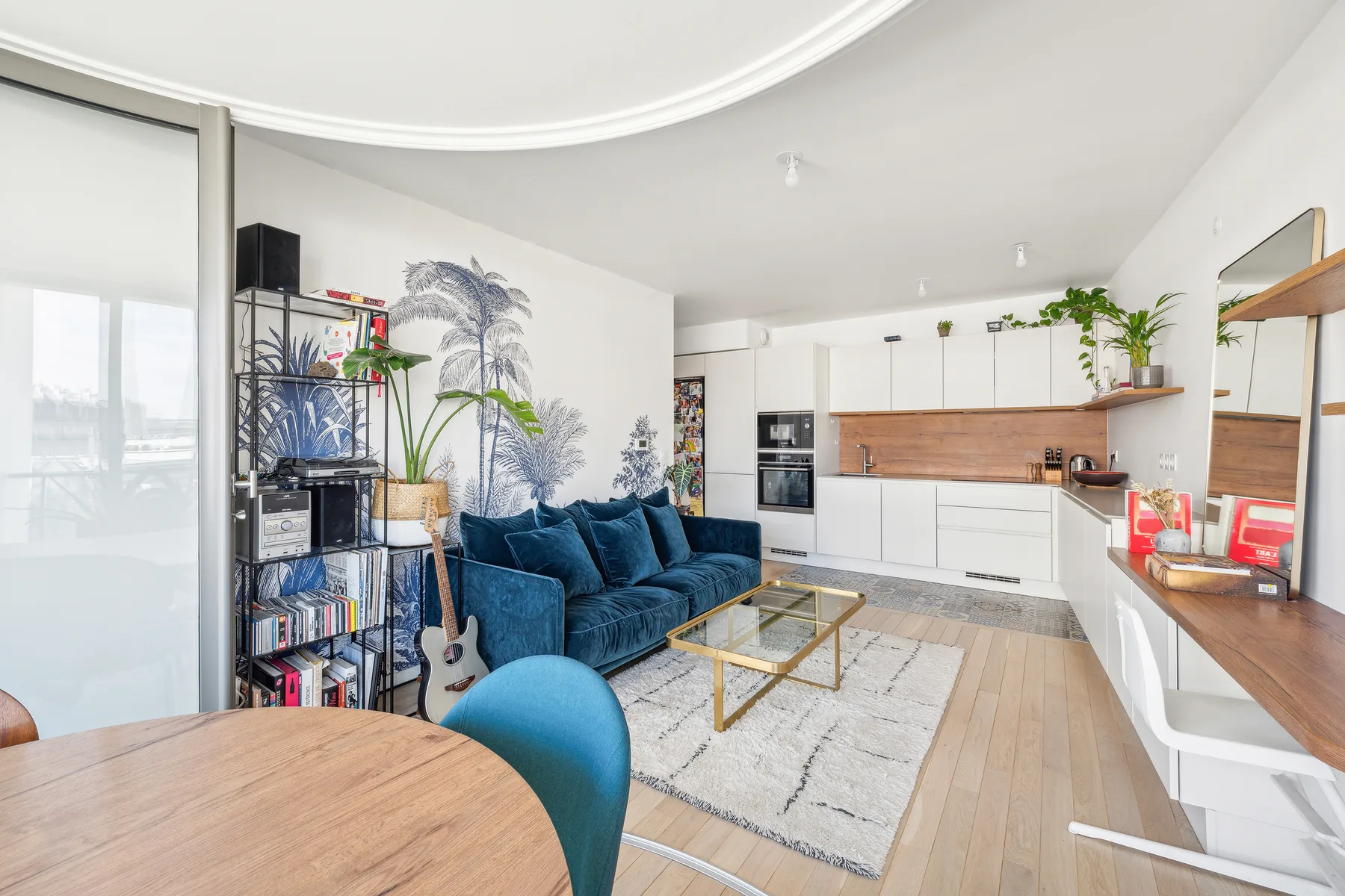 Una sala luminosa se abre a una cocina blanca, con asientos azules y una mesa de centro.