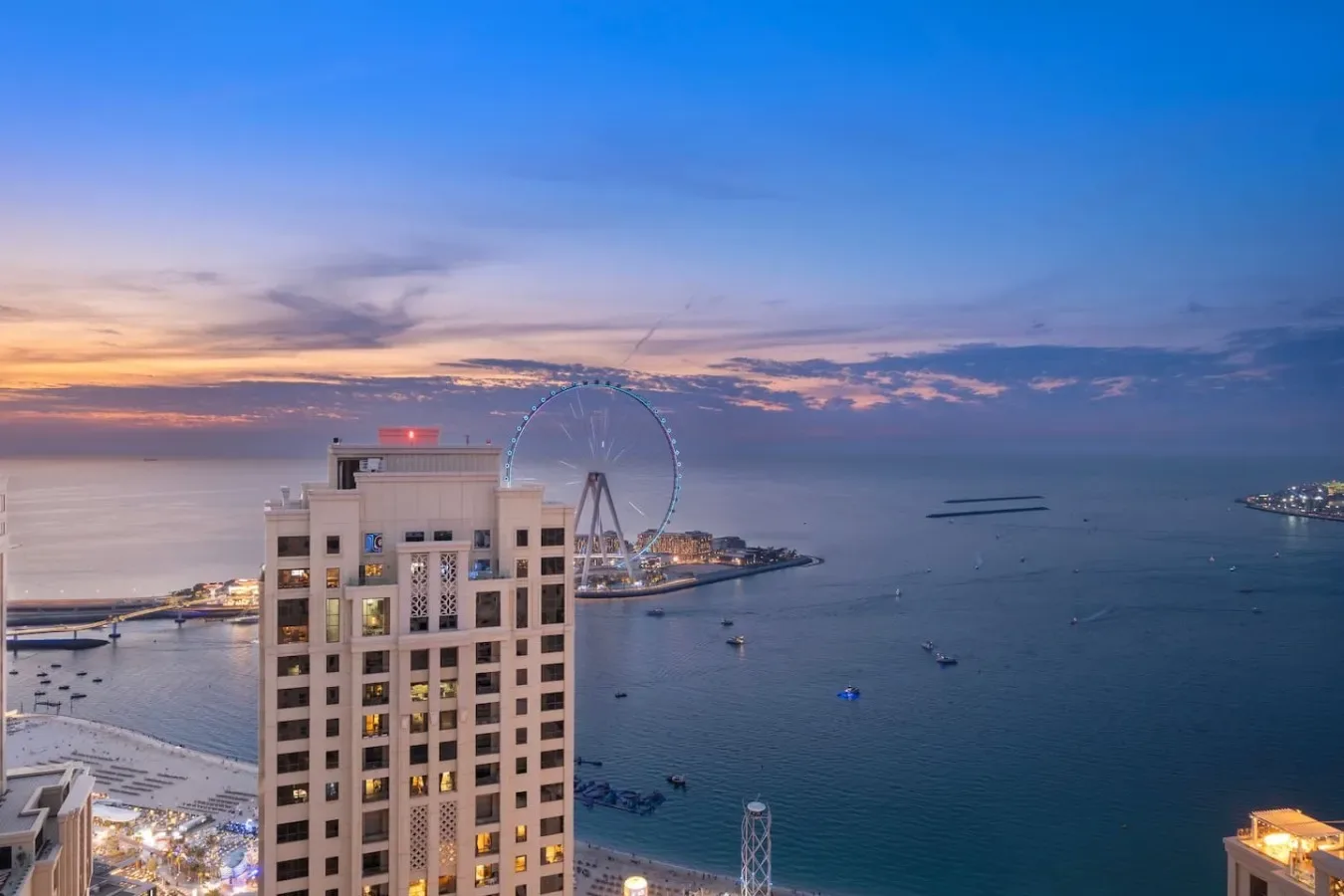 Un edificio alto domina el paseo marítimo y una gran noria al atardecer.