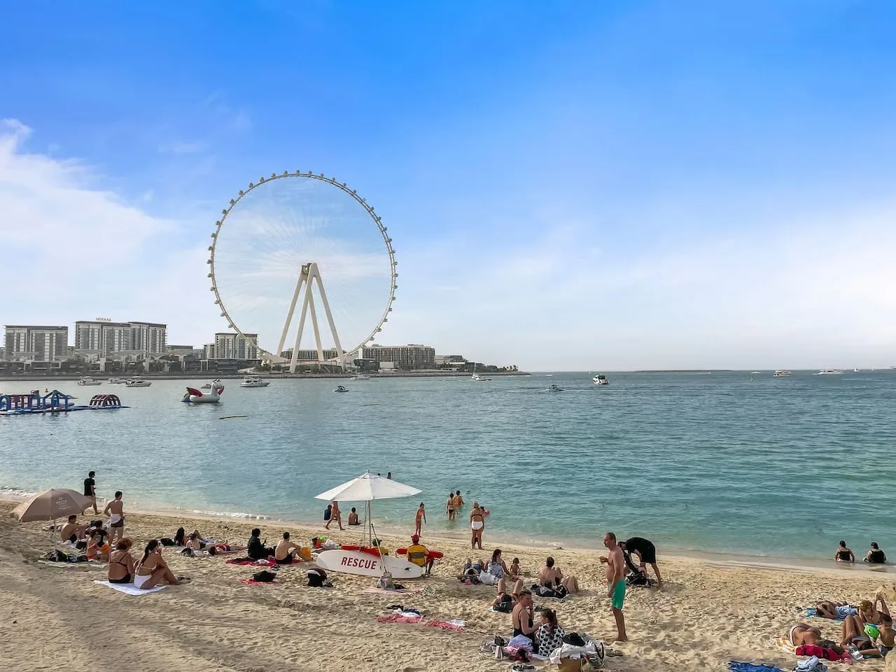Una playa concurrida junto a aguas tranquilas, con una gran noria al otro lado de la bahía.