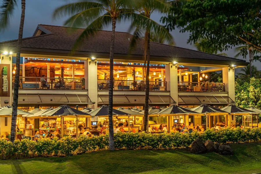 Un restaurante iluminado con calidez y mesas al aire libre, rodeado de palmeras y vegetación.