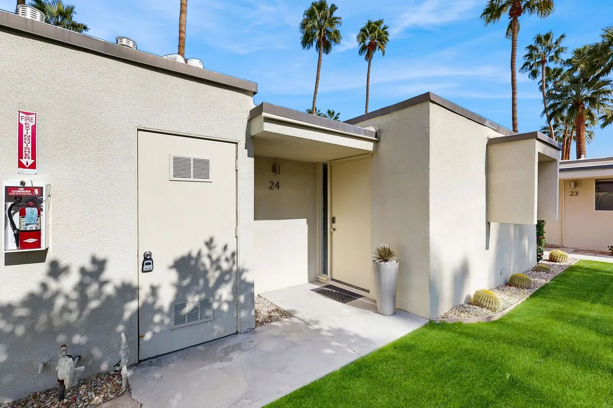 A row of light-colored building entrances opens onto a green lawn beneath palm trees.
