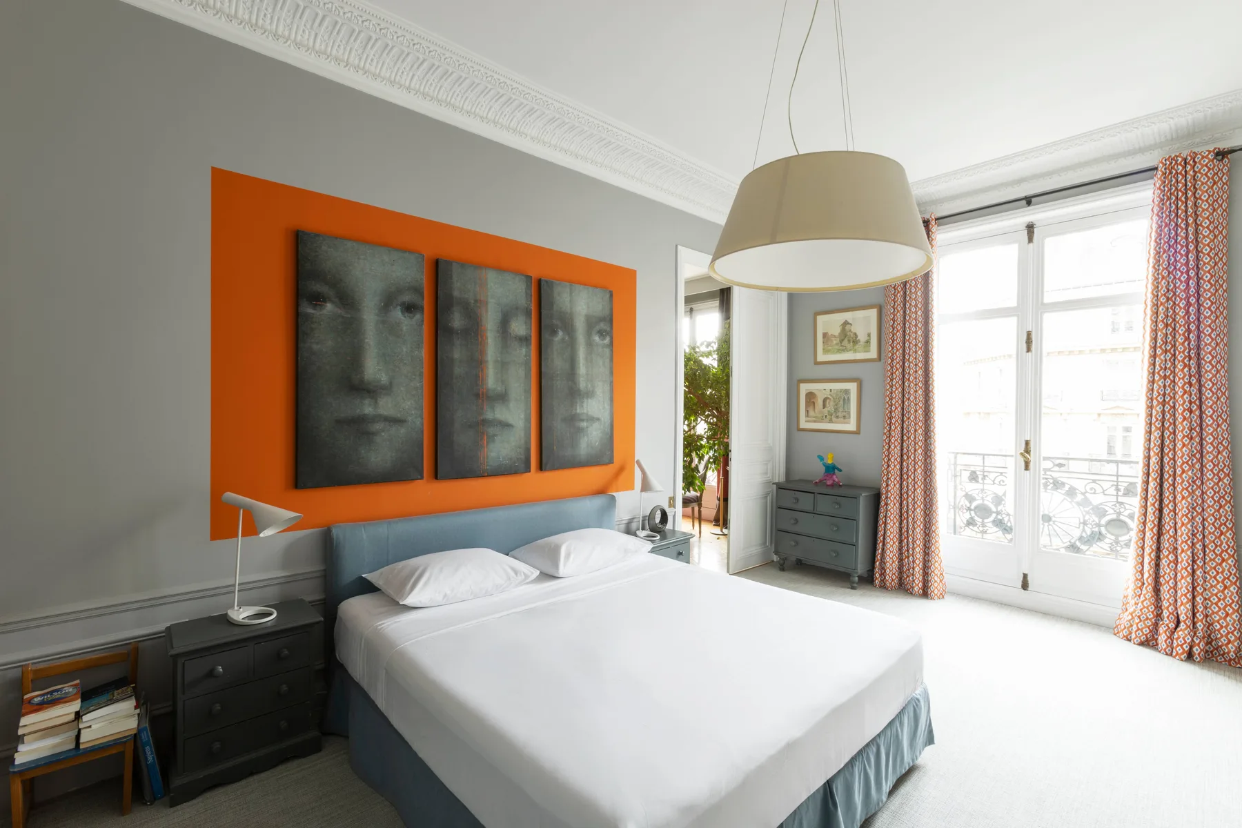 A bright bedroom has a neatly made bed beneath three framed portraits.