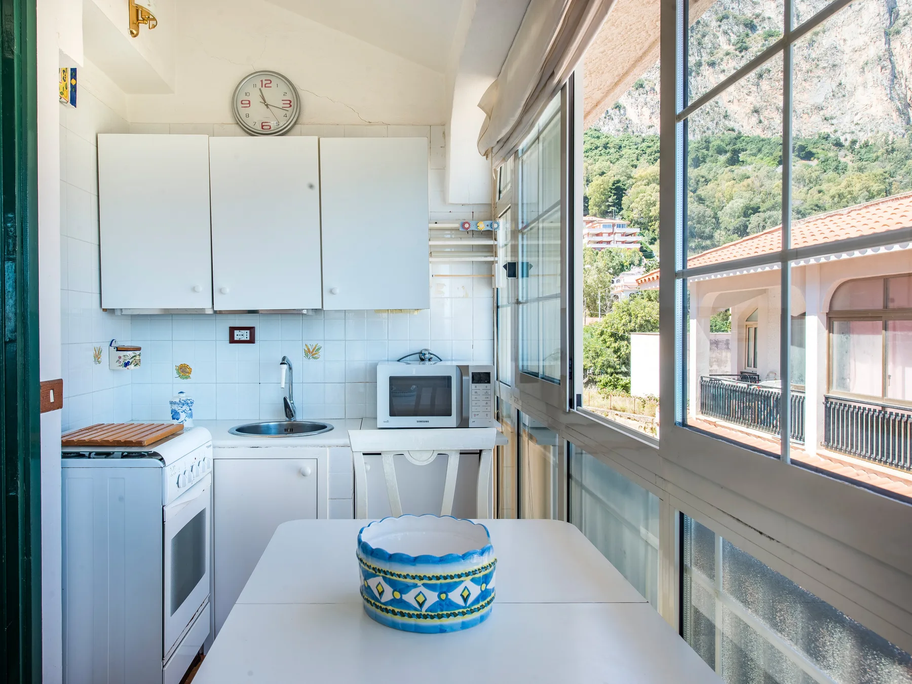 A compact kitchen with a table beside large windows overlooking nearby buildings and a hillside.