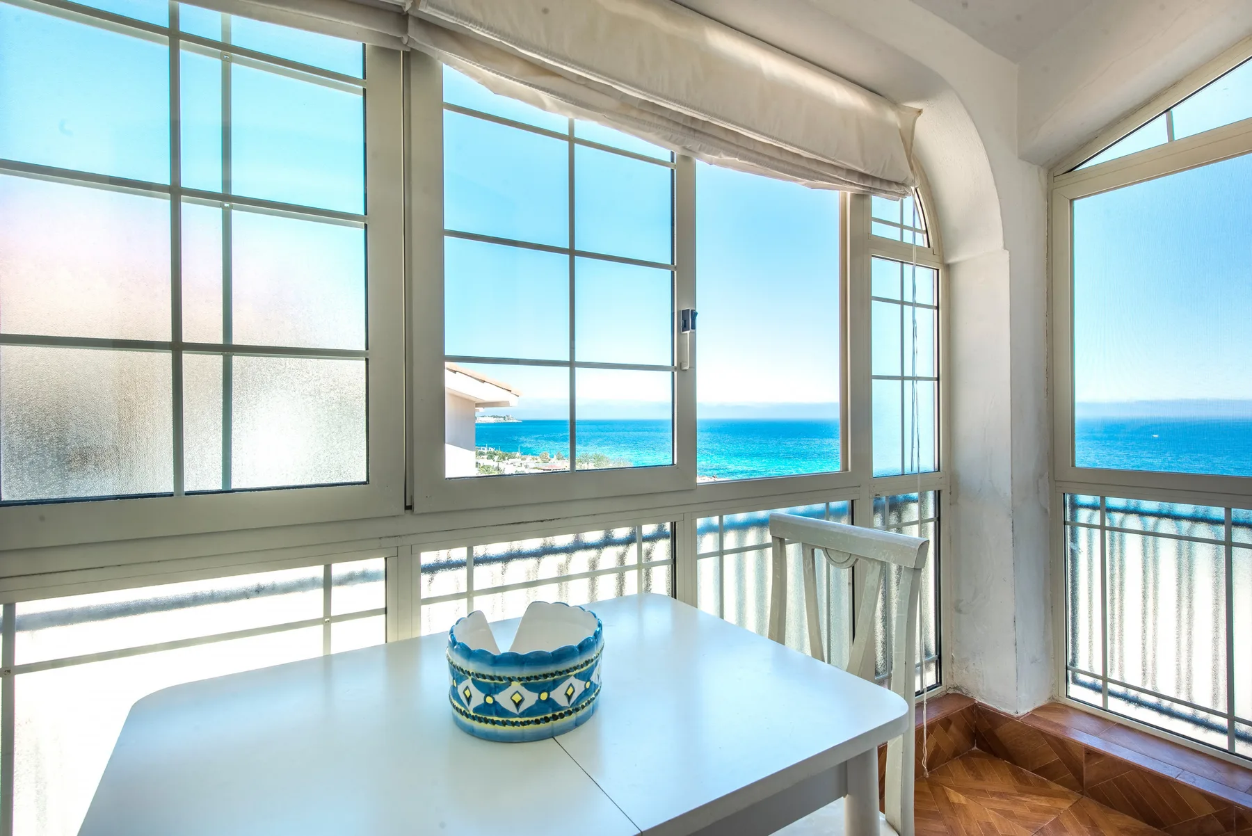 A bright enclosed balcony with a table beside large windows overlooking the sea.