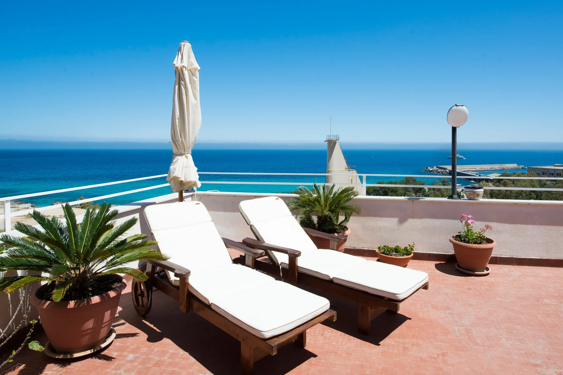 Una terraza soleada junto al mar, con tumbonas y plantas en macetas frente al océano.