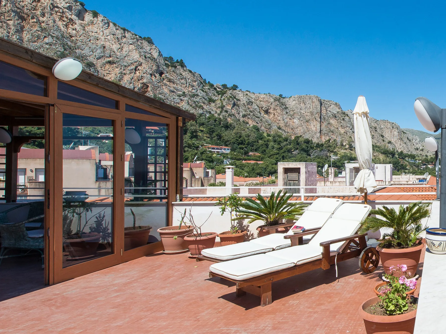 A rooftop terrace with sun loungers and potted plants overlooks a rocky hillside.